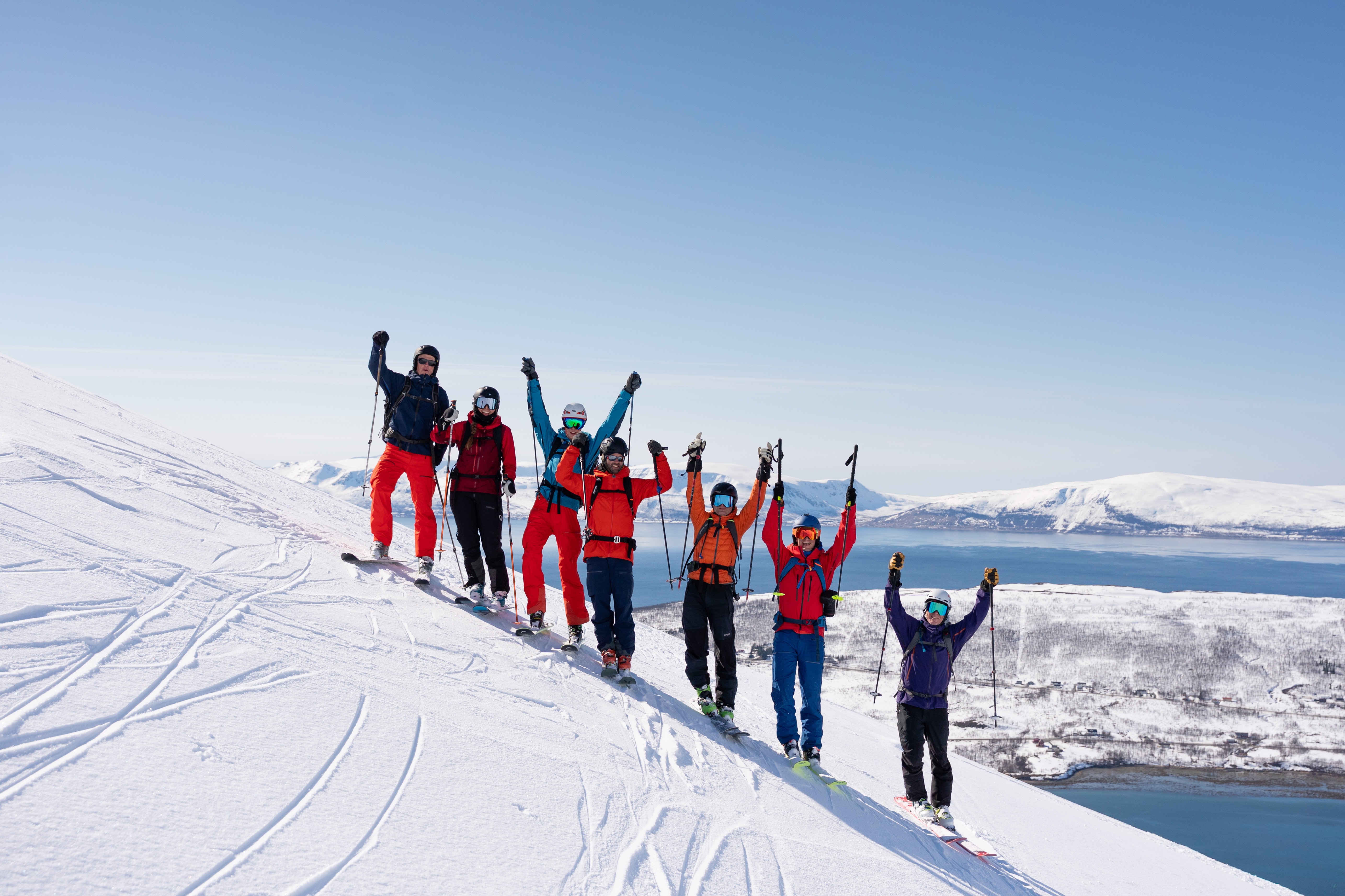 En gruppe skikjørere som står på et fjell med blå himmel og havet bak