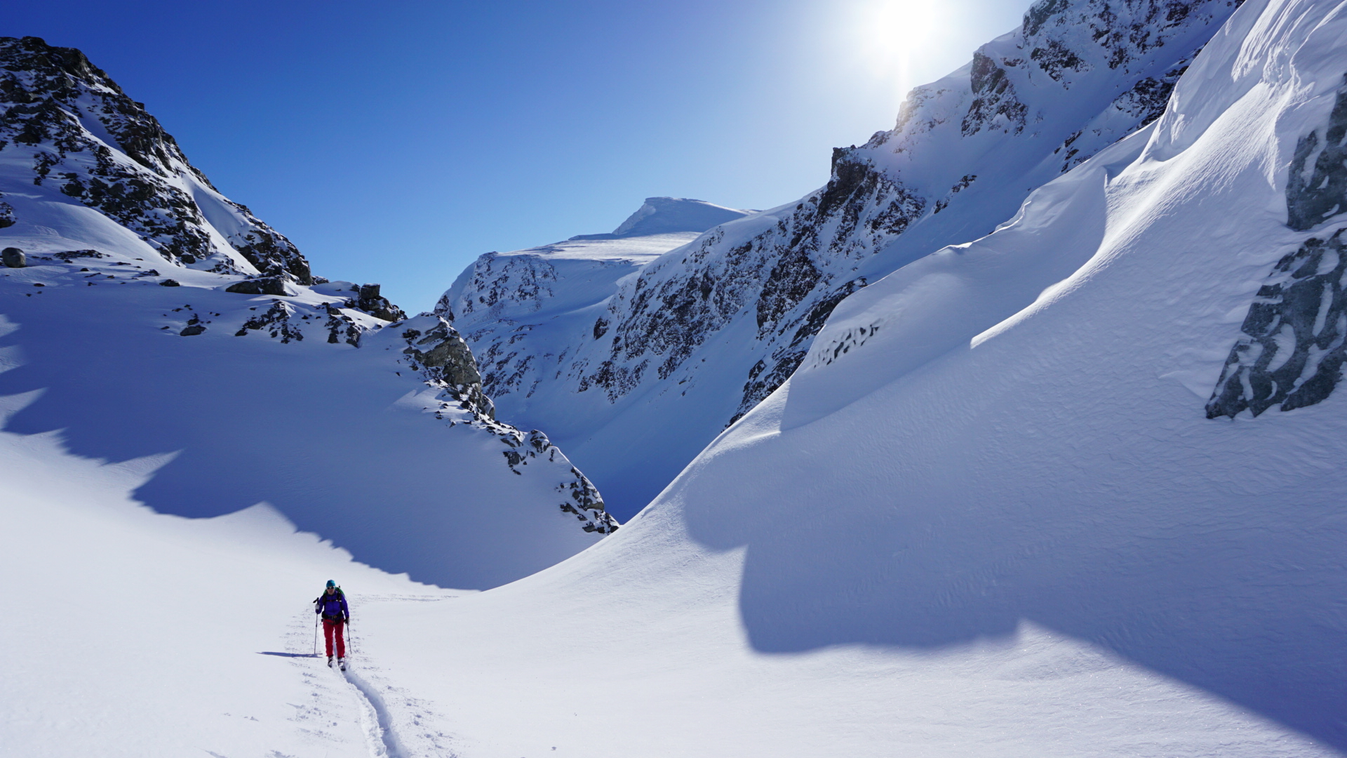 Person som går på ski gjennom en dal full av snø med blå himmel og sol