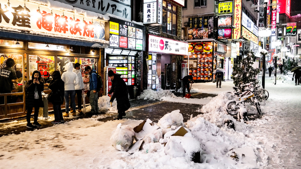 Snow-covered city street with billboards and shop signs, and a few people standing and looking into the shop windows.