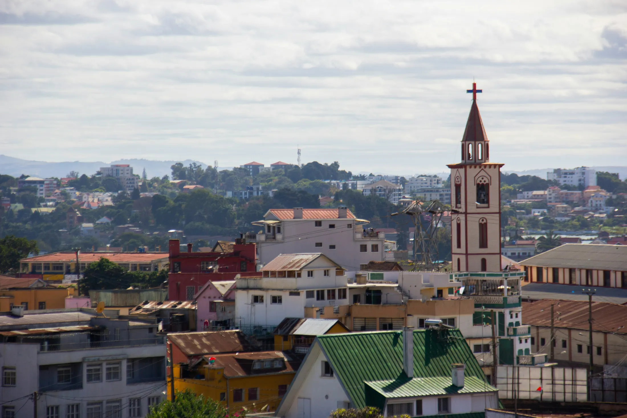 A church steeple with a red cross stands above a dense landscape of colorful city buildings stretching toward distant green hills under a cloudy sky.
