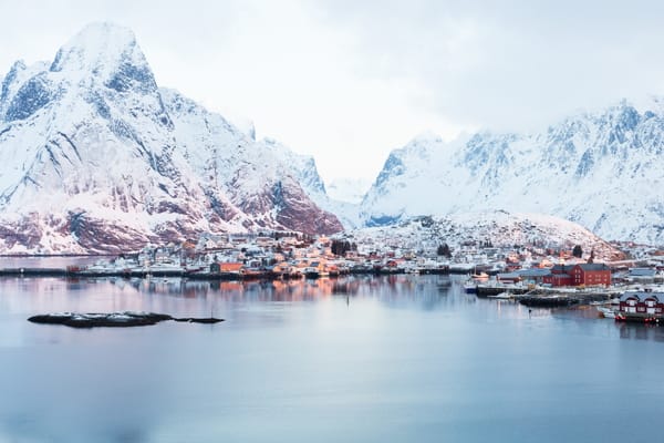 A winter landscape showing a coastal village nestled beneath majestic snow-covered mountains, reflected in calm waters.