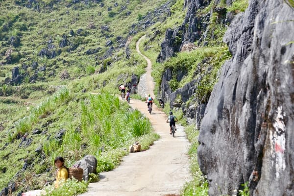 A winding mountain road with cyclists riding uphill, flanked by rocky hillsides and lush green vegetation.