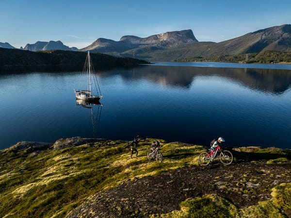 Three mountain bikers pushes their bike up a grassy slope overlooking a calm fjord with a sailboat and mountains in the background.