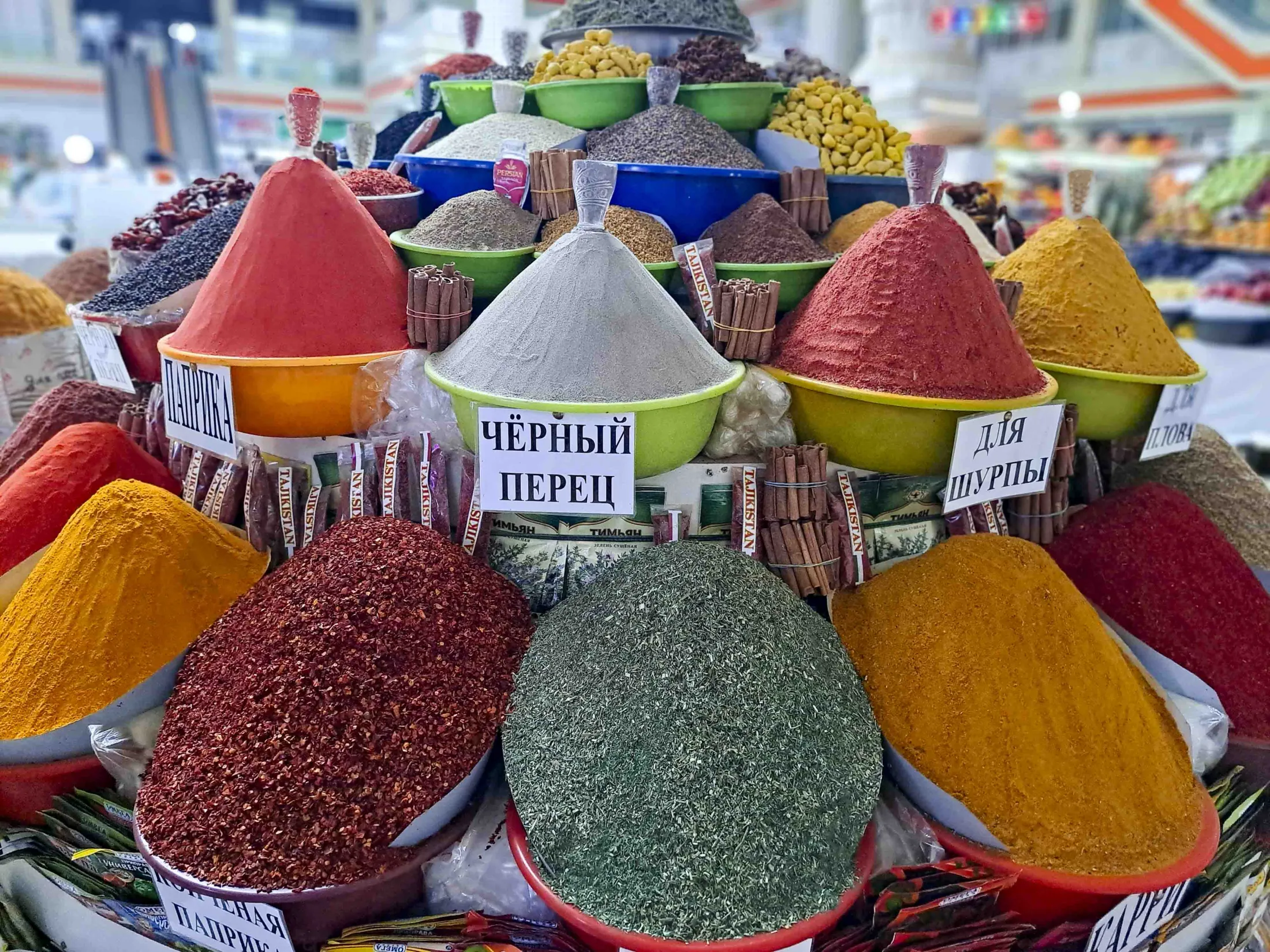 Vibrant mounds of various colorful spices, herbs, and cinnamon sticks are neatly arranged in bowls at a market display with Russian labels.
