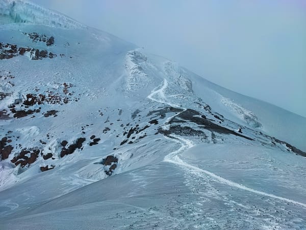 A snowy mountain ridge with a winding trail leading upwards under an overcast sky.