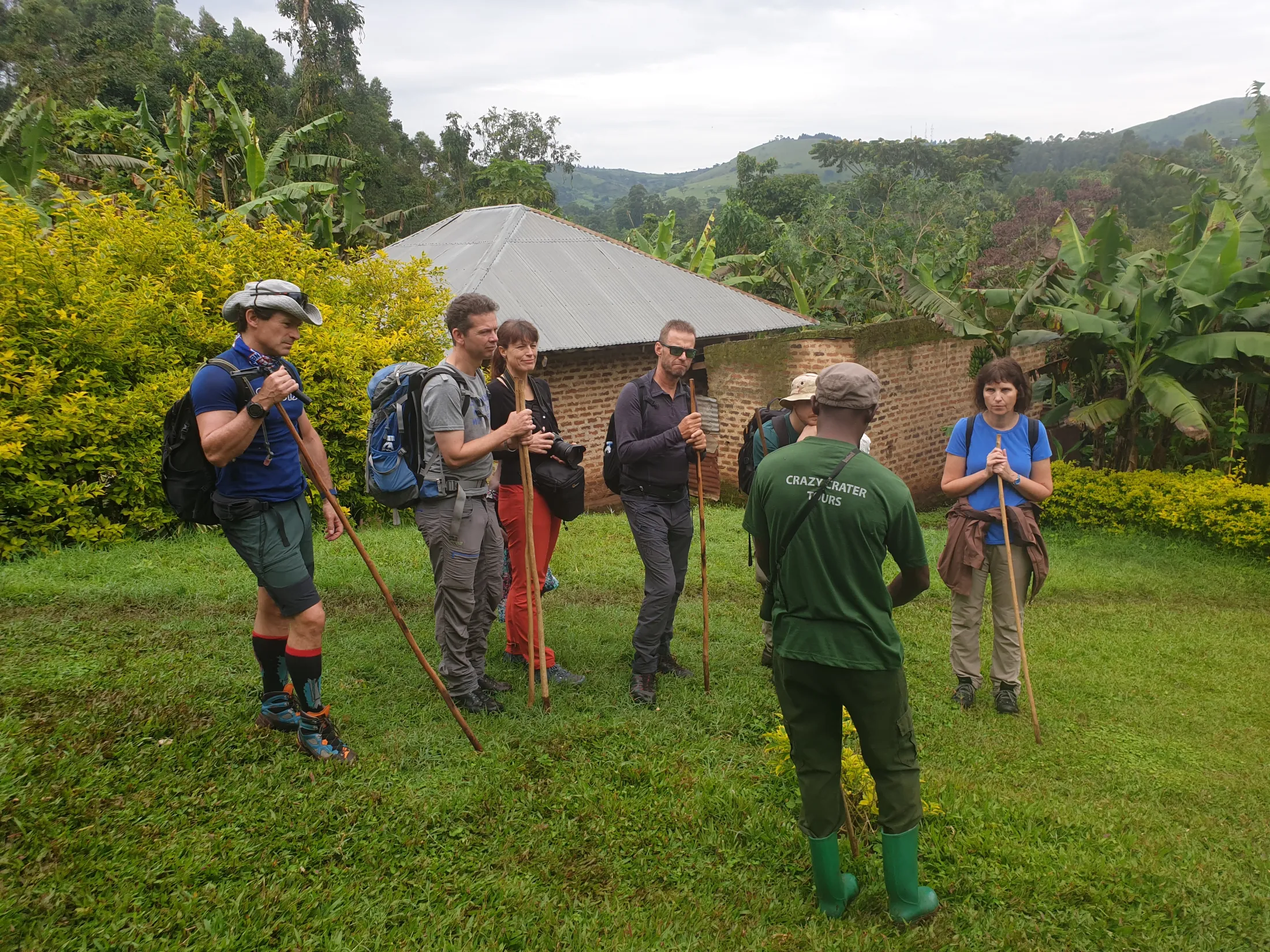 A group of hikers with backpacks standing outdoors on a green lawn near a house and trees.