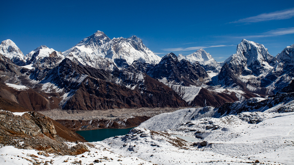 A stunning panoramic high-altitude view shows a deep teal glacial lake nestled below a vast network of rugged, snow-capped mountain ranges and massive rocky peaks under a brilliant, clear blue sky.