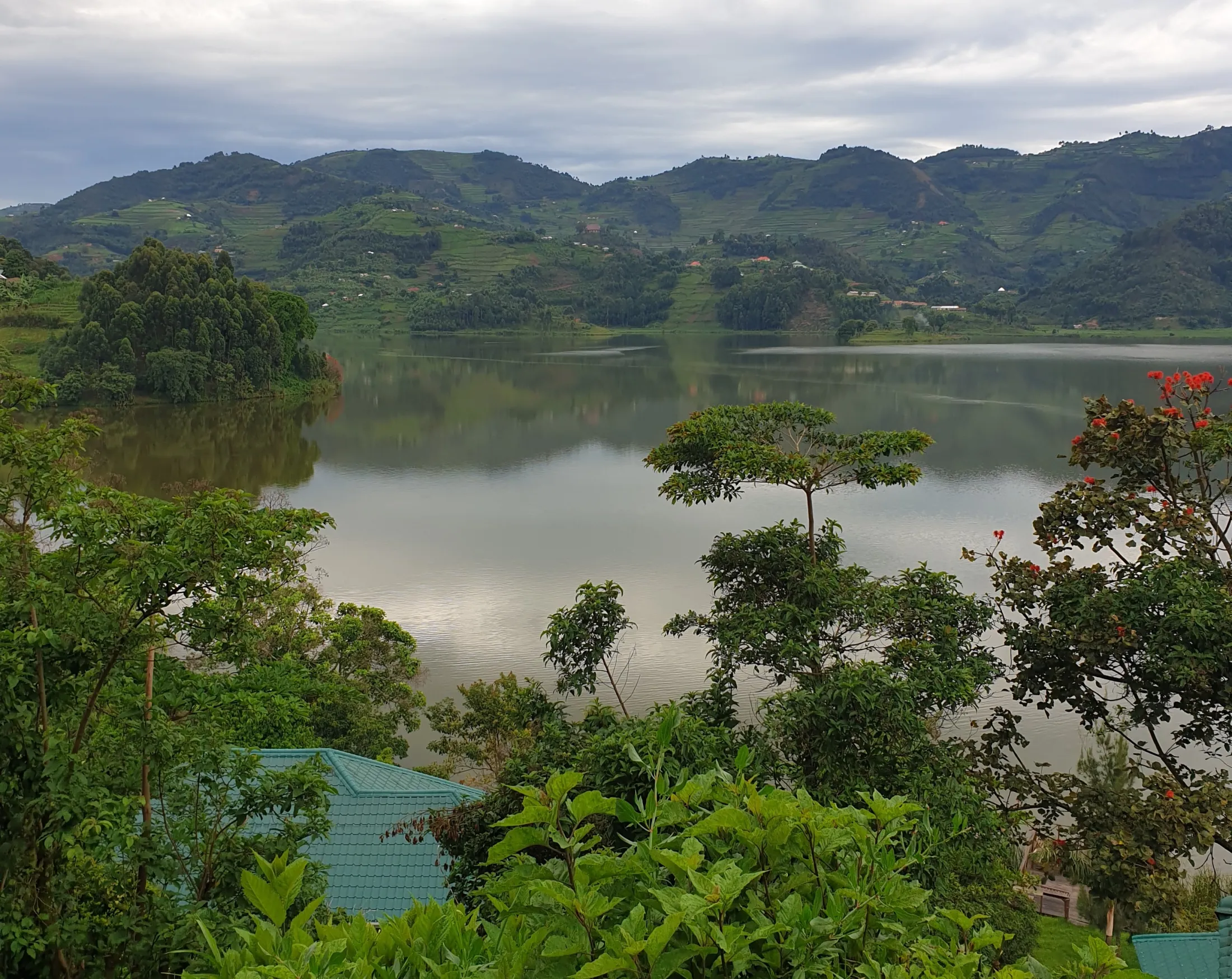 A serene landscape with a lake reflecting overcast sky, surrounded by lush green trees and distant hills under a cloudy sky.