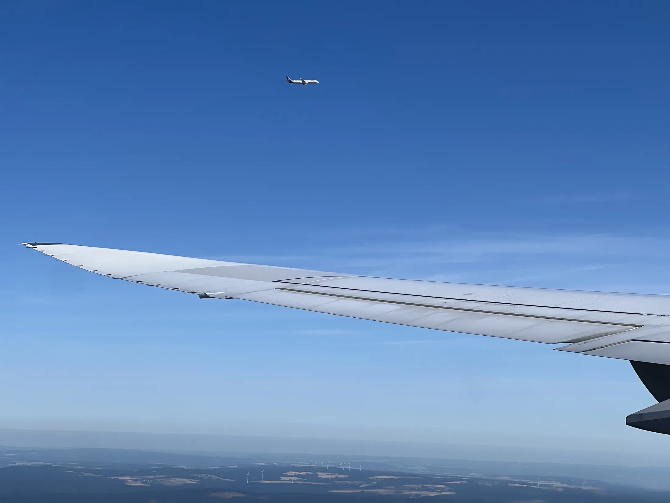 The wing of an airplane extends into the foreground against a clear blue sky, while another commercial aircraft flies parallel at a higher altitude in the distance.