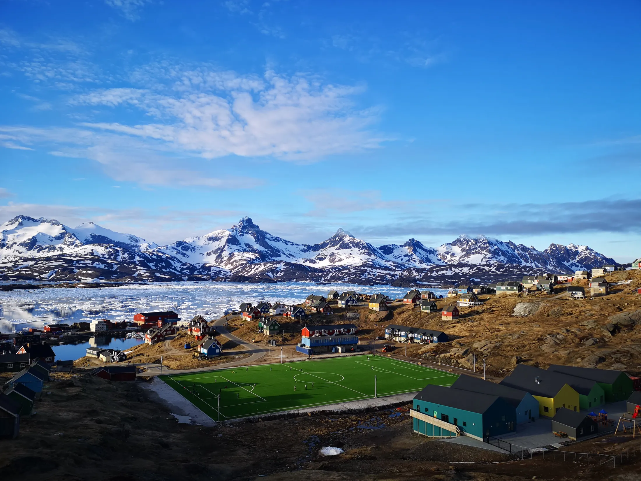 A green soccer field sits in the middle of a colorful Greenlandic settlement, with an ice-filled fjord and sharp, snow-covered mountains under a blue sky in the background.