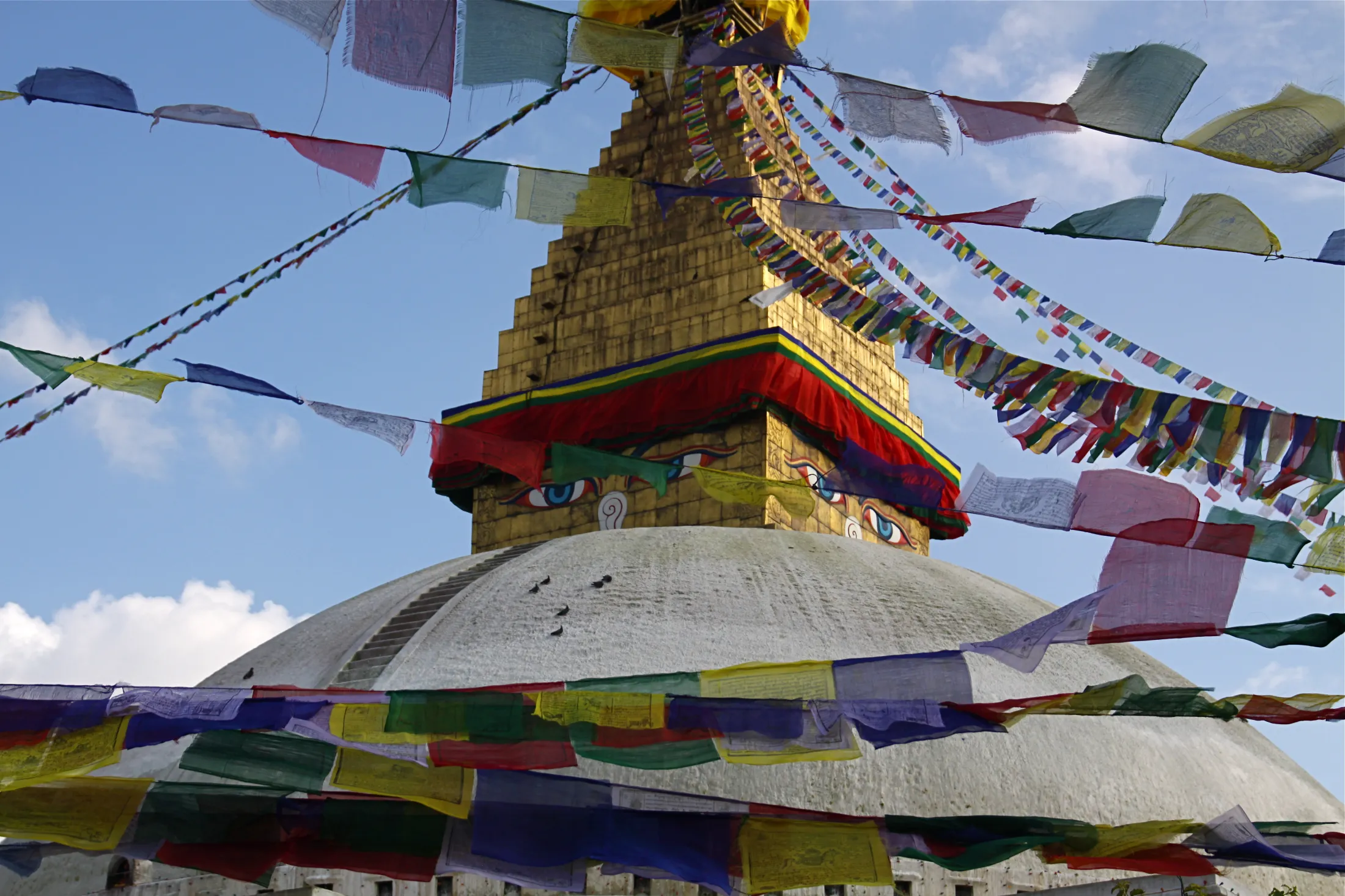 A close-up view of a large white stupa with the painted eyes of the Buddha on its golden spire, crisscrossed by numerous colorful Buddhist prayer flags under a partly cloudy blue sky.