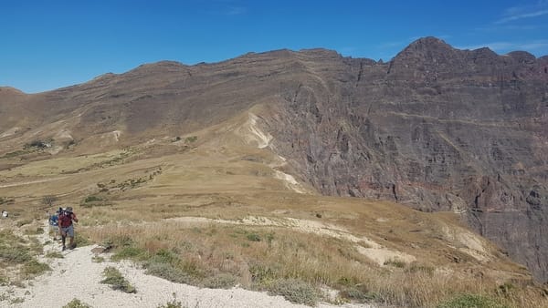 A narrow dirt path traces along a barren, light-colored ridge leading toward massive, dark volcanic mountain slopes under a clear blue sky.