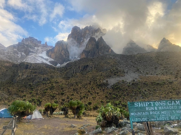 A sign for Shipton's Camp stands at a high-altitude campsite on Mount Kenya, with tents pitched below dramatic, misty rock peaks rising into the clouds.
