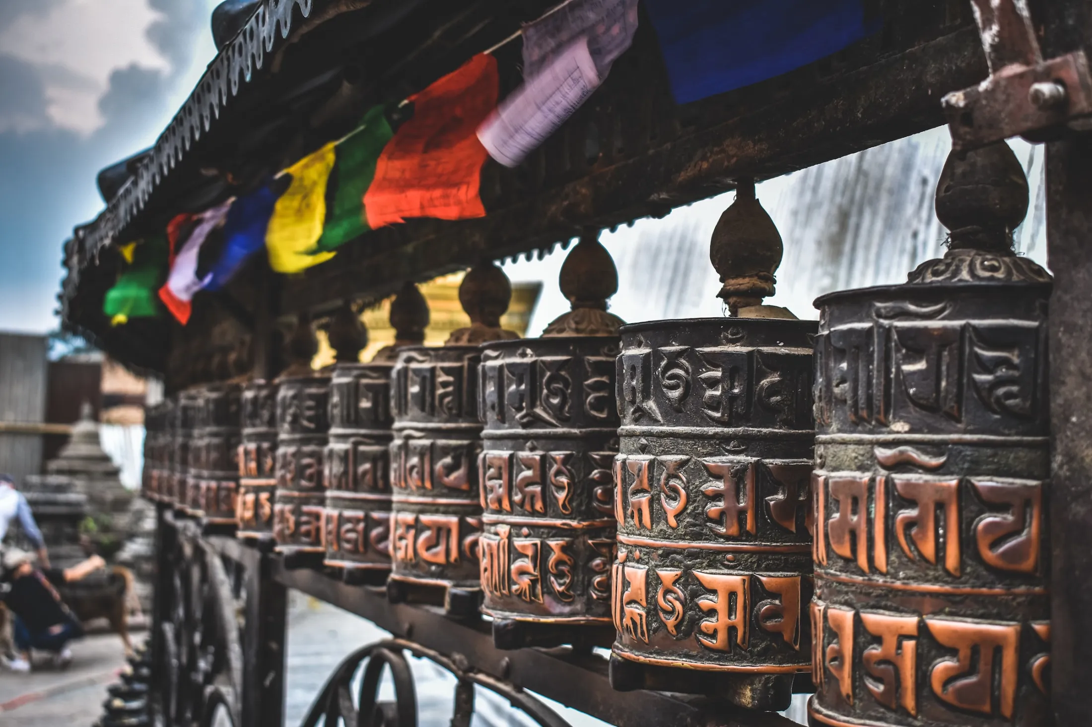A row of weathered, embossed metal Buddhist prayer wheels lines a structure, with colorful prayer flags hanging above them under a cloudy sky.
