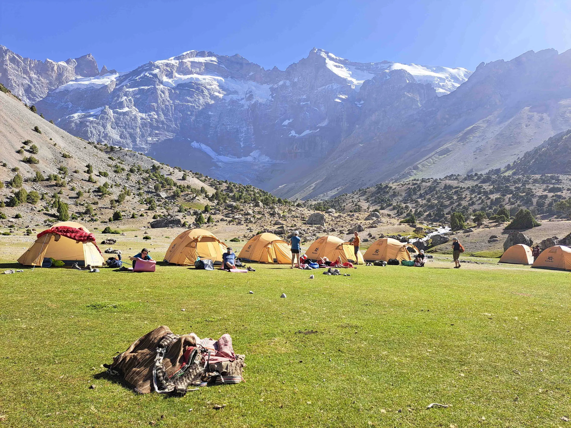 Several orange tents are pitched on a lush green grassy field where people are relaxing, set against a backdrop of towering, snow-capped mountains under a bright sky.