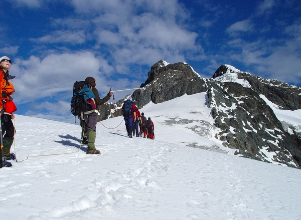 A team of mountaineers roped together walks across a steep, snow-covered glacier towards a towering rocky peak under a partly cloudy blue sky.