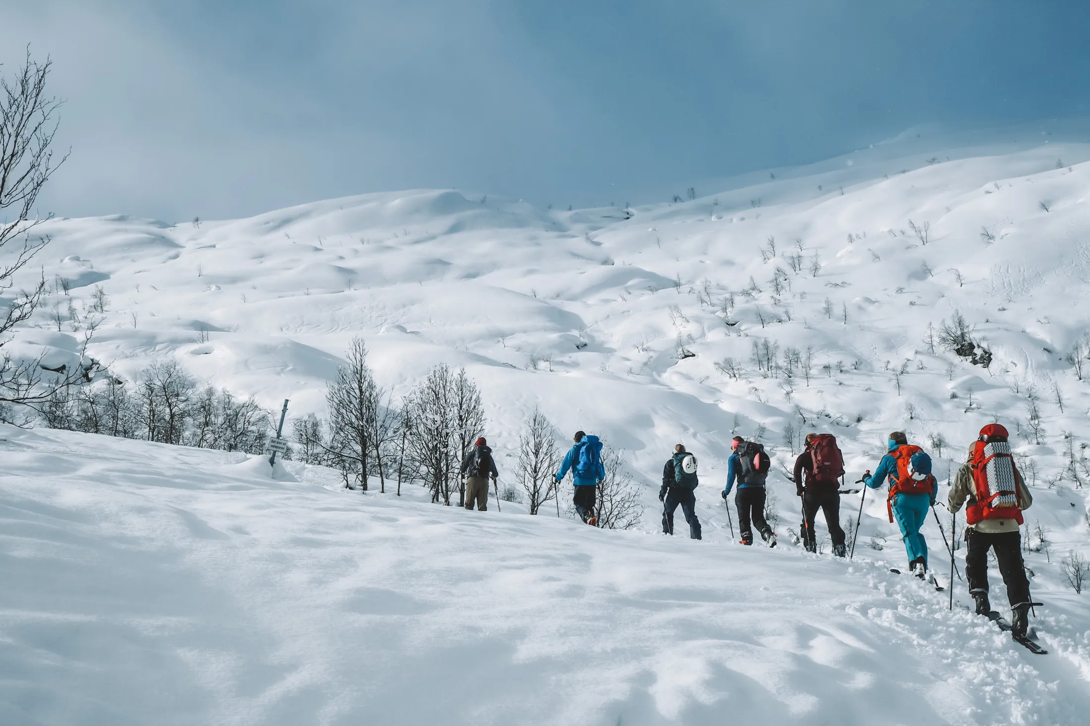 A group of people on skis travels in a long line across a snow-covered mountain landscape under a blue sky.