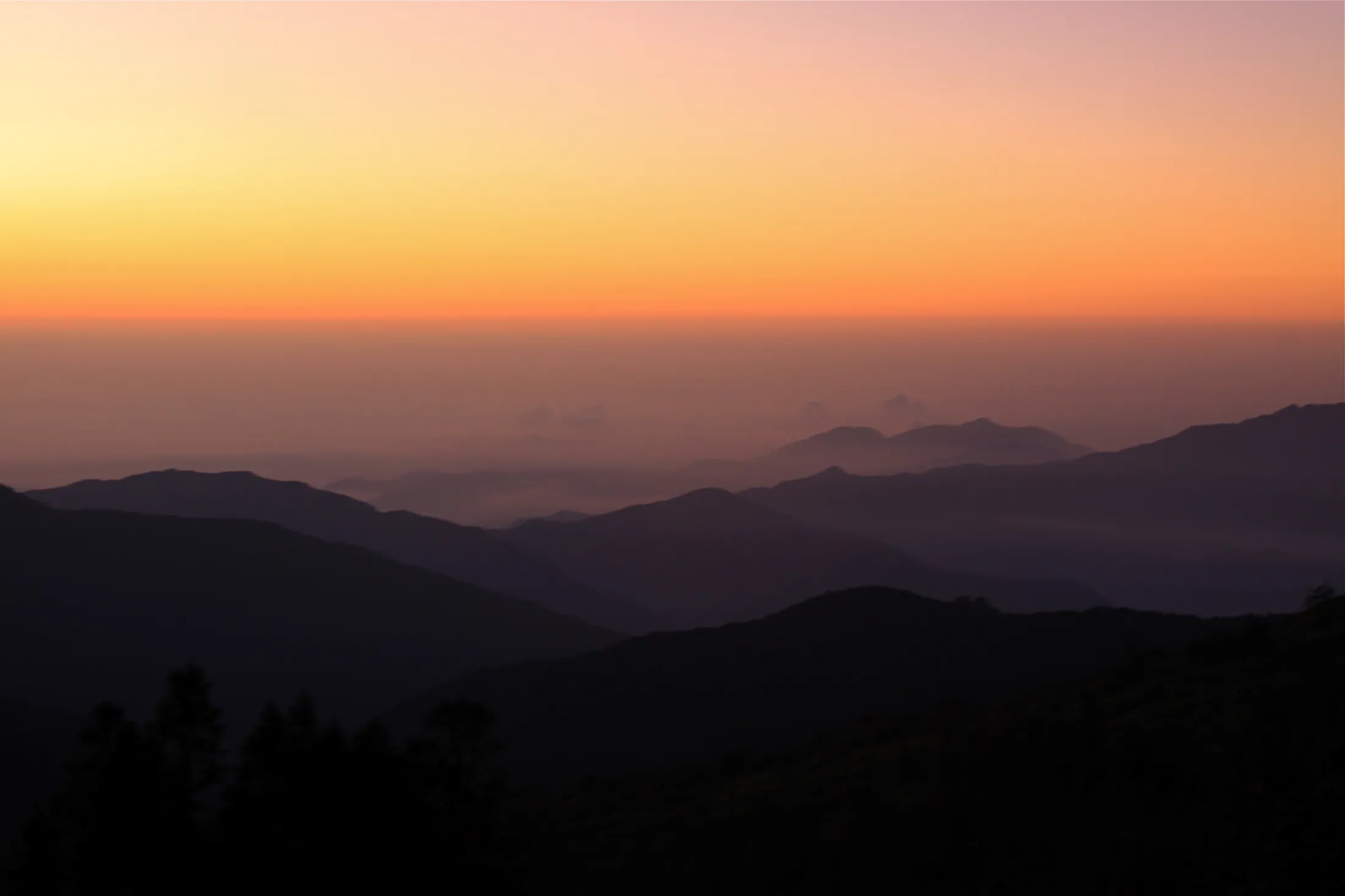 Layered silhouettes of rolling mountain ridges stretch across a misty valley under a vibrant gradient sky shifting from deep orange to soft yellow at dawn.