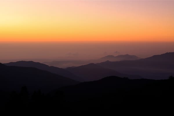 Layered silhouettes of rolling mountain ridges stretch across a misty valley under a vibrant gradient sky shifting from deep orange to soft yellow at dawn.