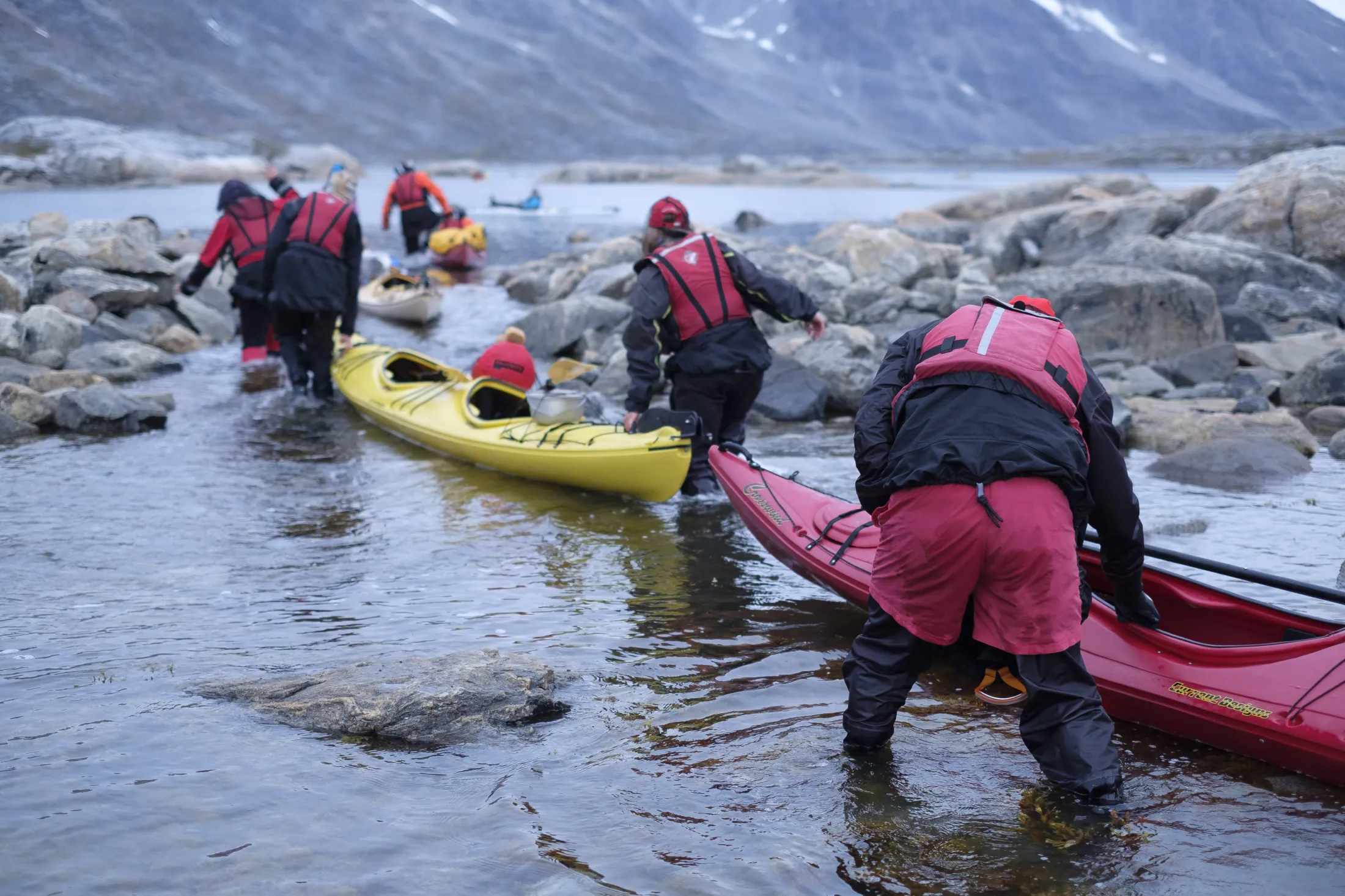 A group of kayakers in drysuits and life jackets work together to move their kayaks through a shallow, rocky channel.