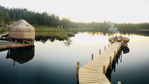 A wooden pier extends over a calm lake towards a seating area, with a yurt-like building on the left bank.