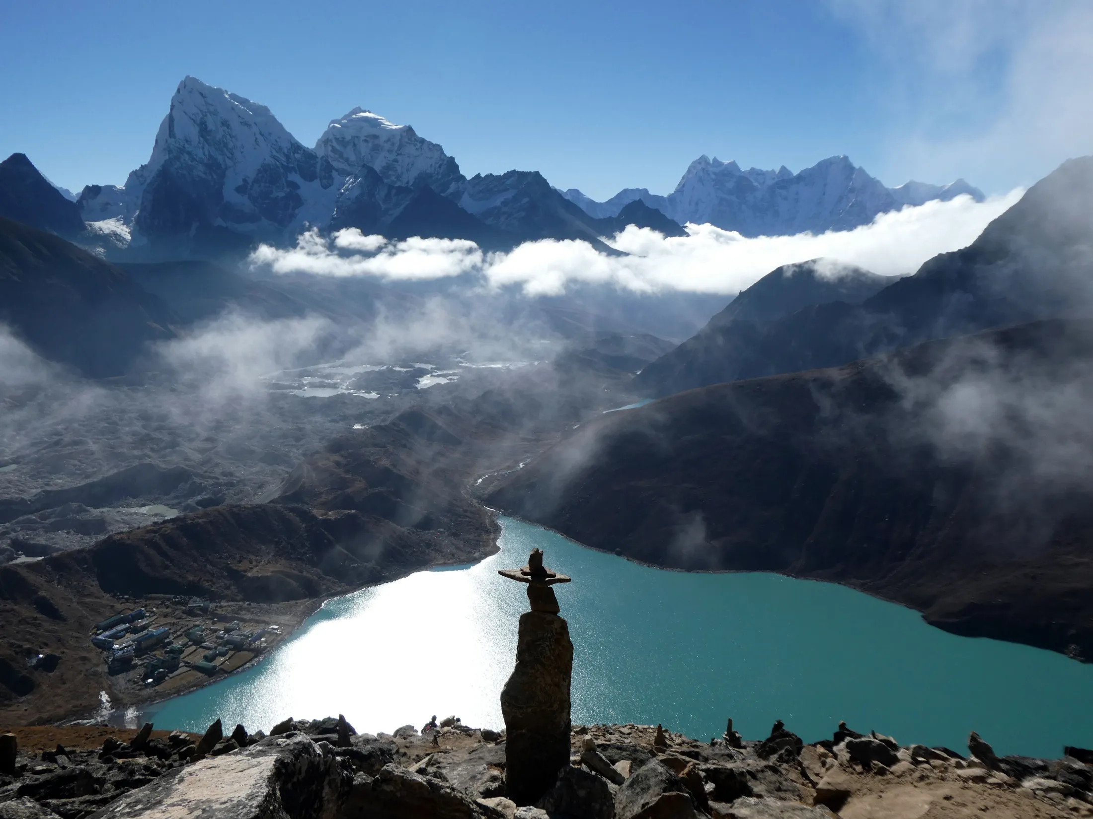 A stone cairn stands on a rocky peak in the close foreground, overlooking a brilliant turquoise alpine lake with a glint of sunlight reflecting off the water, surrounded by swirling low clouds and a majestic backdrop of snow-capped mountain peaks.