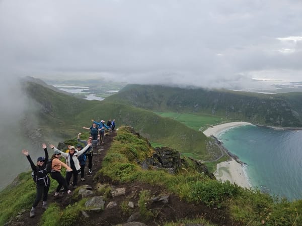 A group of hikers standing on a ridge with hands raised, overlooking a beach and sea under an overcast sky.