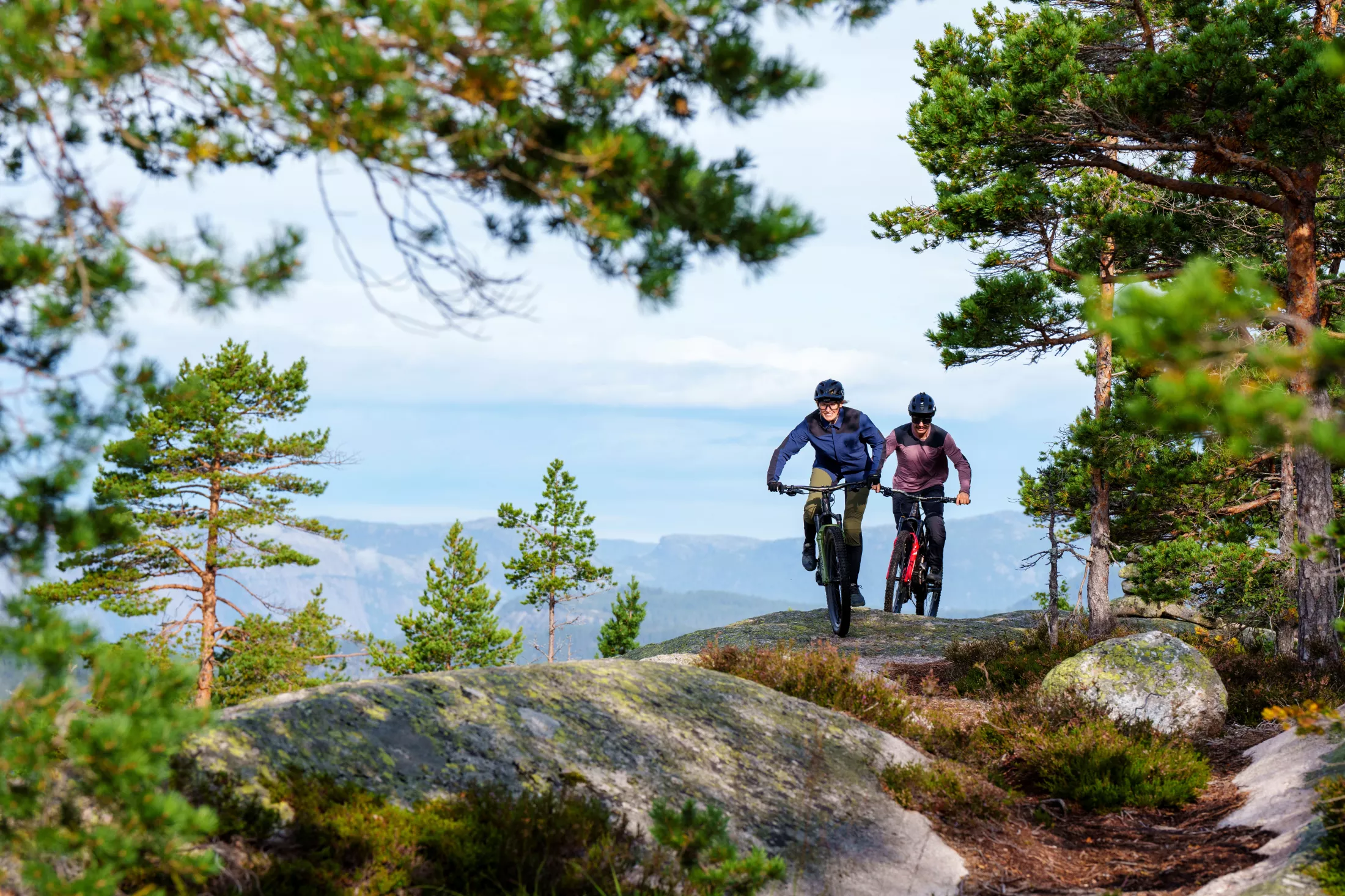 Two mountain bikers cycling along a scenic forest trail with mountains in the background.