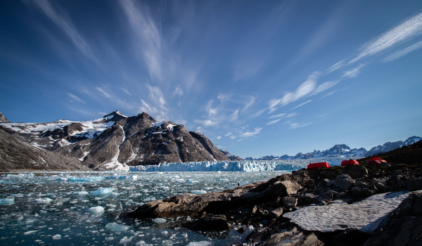 A campsite with red tents sits on a rocky ridge overlooking an ice-filled fjord, with a massive glacier front and majestic, partly snow-covered mountains under a dramatic sky in the background.