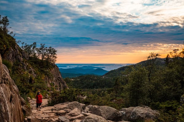 A hiker wearing a red jacket walks along a rocky mountain path at sunset, overlooking a forested valley.