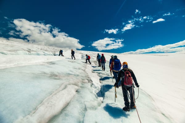 A group of hikers with backpacks and helmets walking on a snowy glacier under a blue sky.