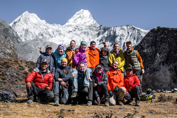 A group of thirteen diverse hikers wearing colorful winter jackets smile and pose closely together for a group photo on a dry, grassy flat, with massive snow-capped mountains under a clear sky behind them.