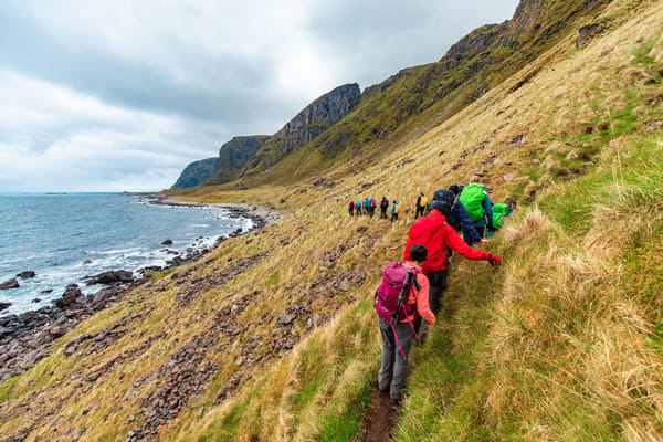 A group of hikers in colorful jackets walking on a steep grassy slope along a rocky coastline under an overcast sky.