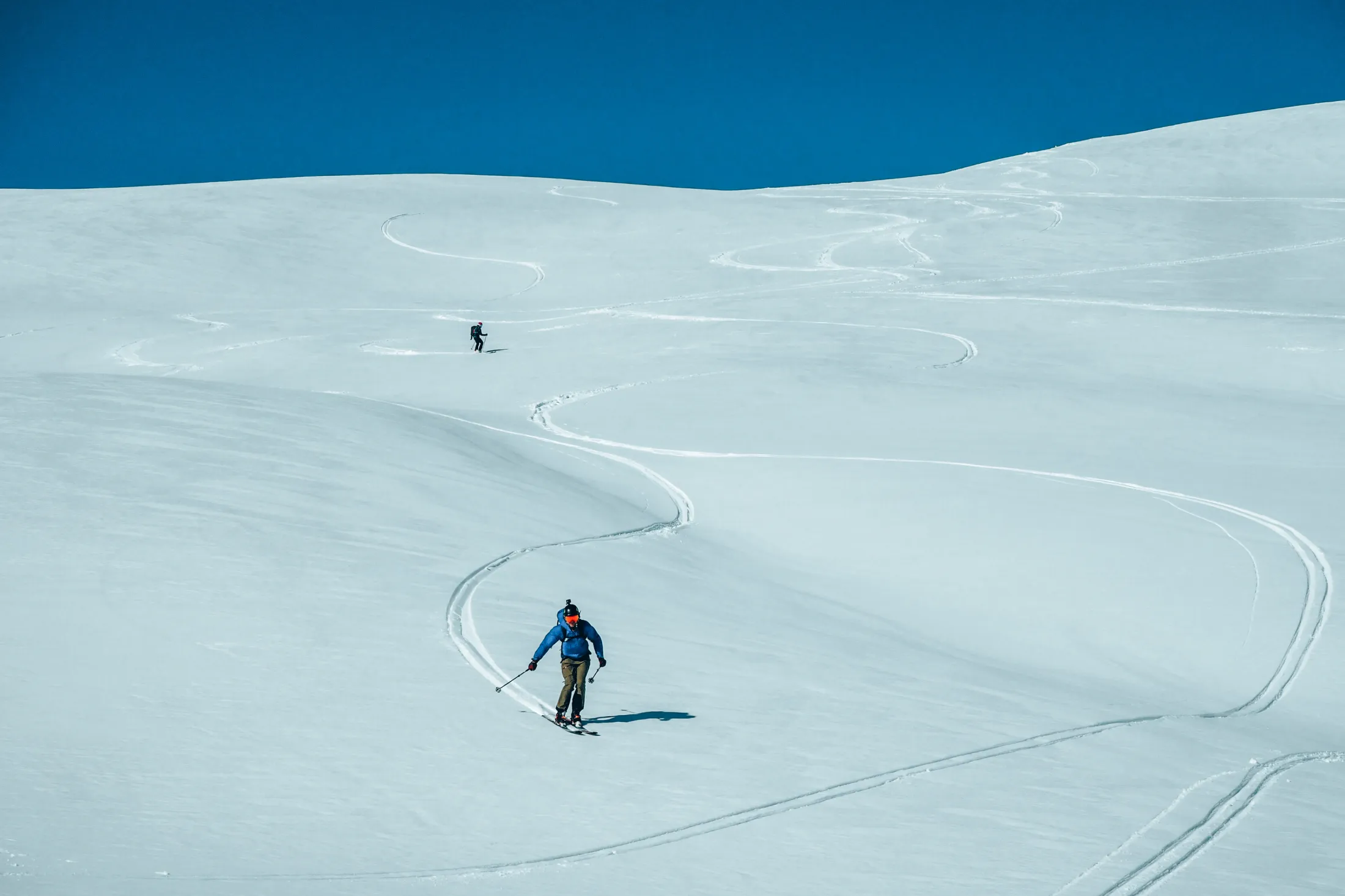 Two skiers carve their way down an untouched, snow-covered mountainside, leaving long tracks in the snow under a clear blue sky.