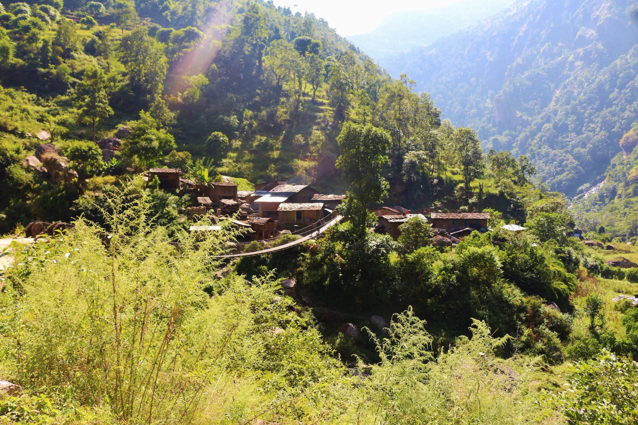 A small cluster of stone buildings sits on a sun-drenched, terraced green hillside surrounded by dense vegetation in a mountain valley.