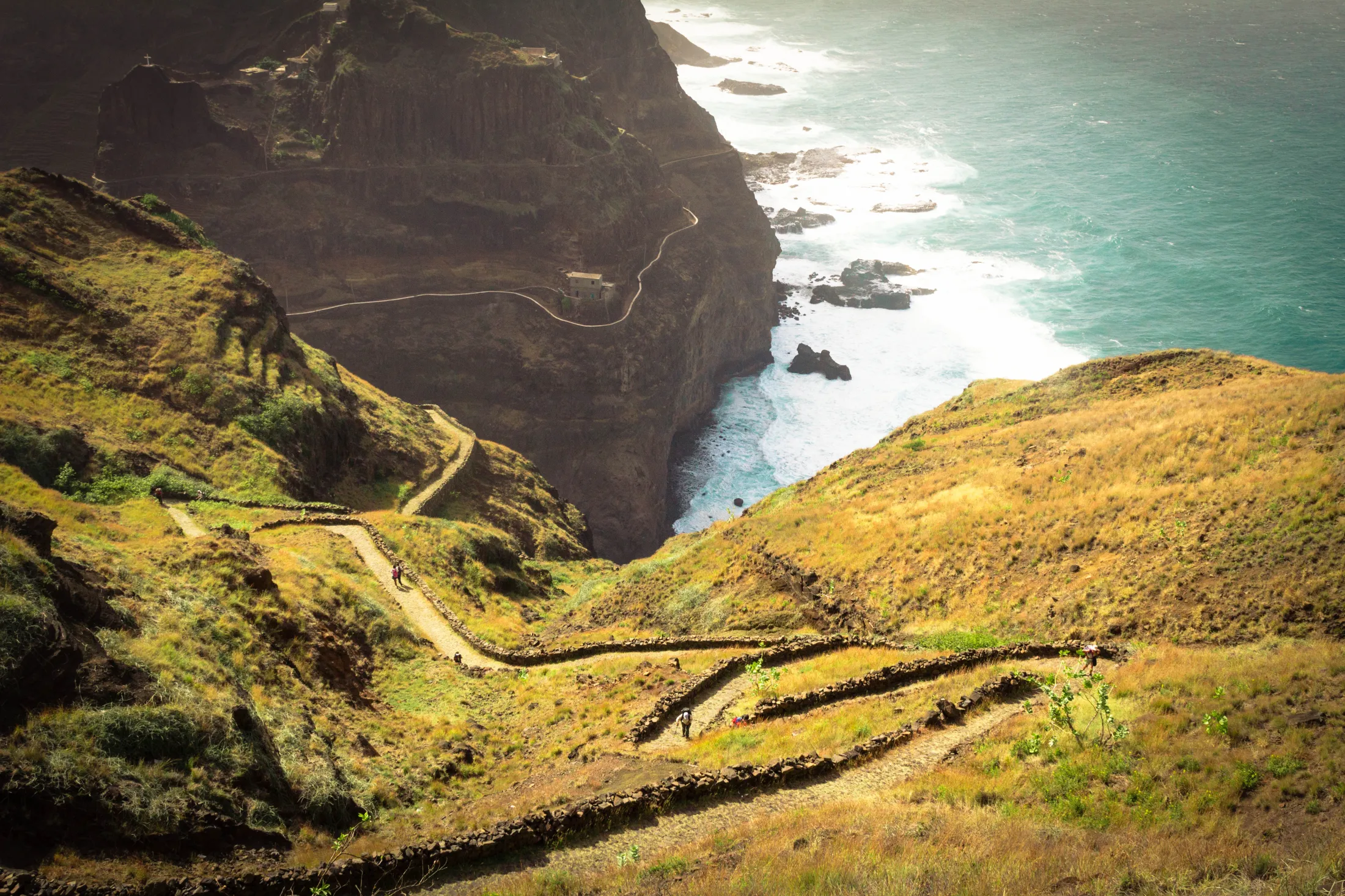 A winding cobblestone hiking path snakes down a steep, grassy valley toward a dramatic coastal cliff side where ocean waves crash against the rocks below.
