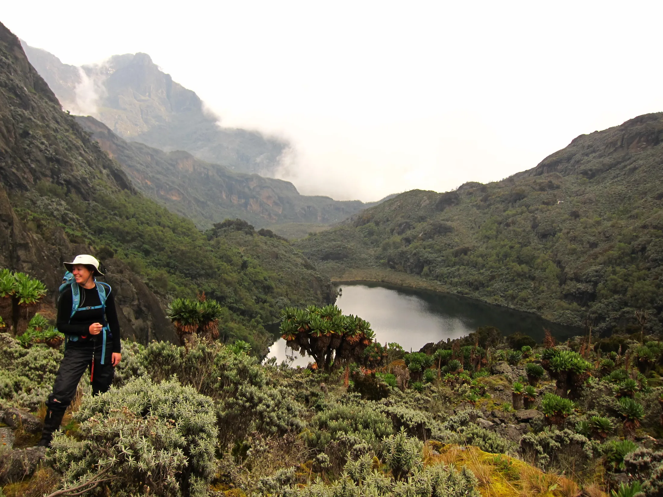 A smiling hiker stands on a vegetated slope overlooking a calm mountain lake nestled within a deep valley under misty peaks.