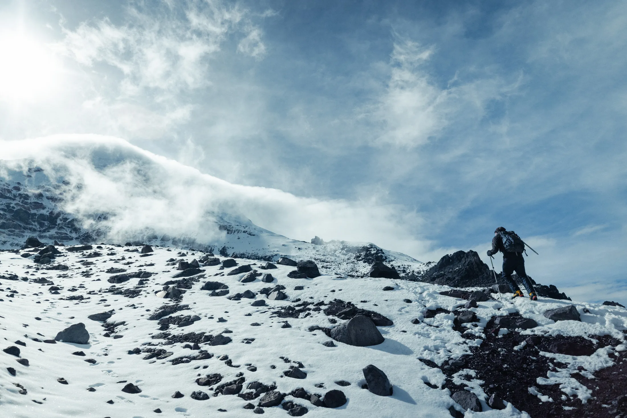 A hiker ascends a snow-covered, rocky mountain slope under a blue sky with cloud layers.