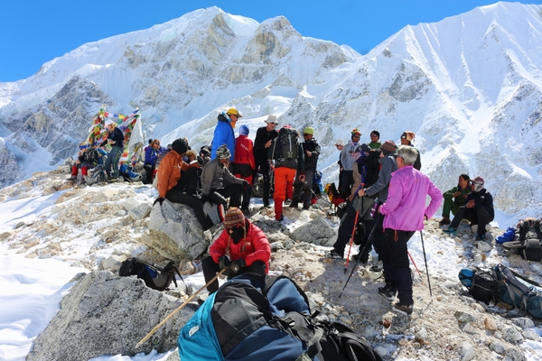 A large group of mountaineers and hikers rest on a rocky, snow-dusted ridge beneath a towering, bright white glacial mountain wall.