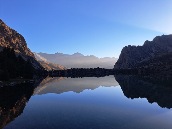 A calm mountain lake perfectly mirrors the surrounding rugged peaks and clear blue sky in a symmetrical reflection.