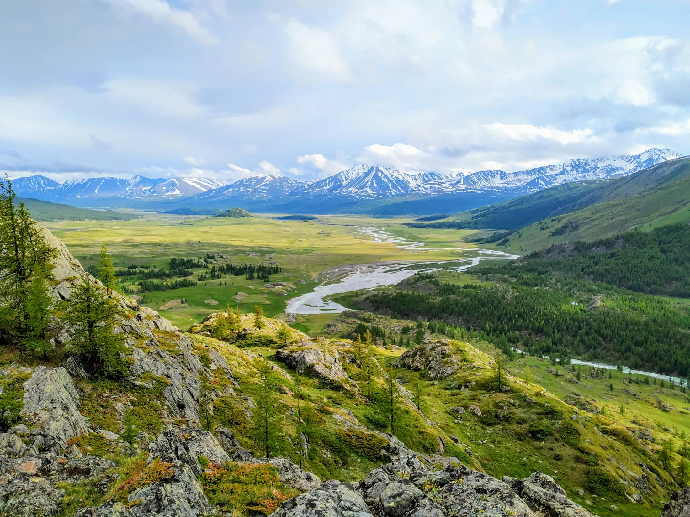 A wide landscape shot of a lush green valley in Mongolia with a winding river flowing through it, framed by rugged, rocky terrain in the foreground and snow-capped mountains under a cloudy sky in the background.