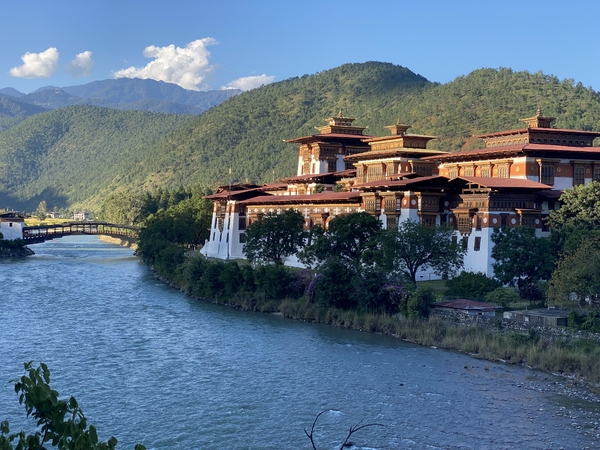 The grand Punakha Dzong fortress situated along a blue river with a wooden cantilever bridge and lush green hills in the background.