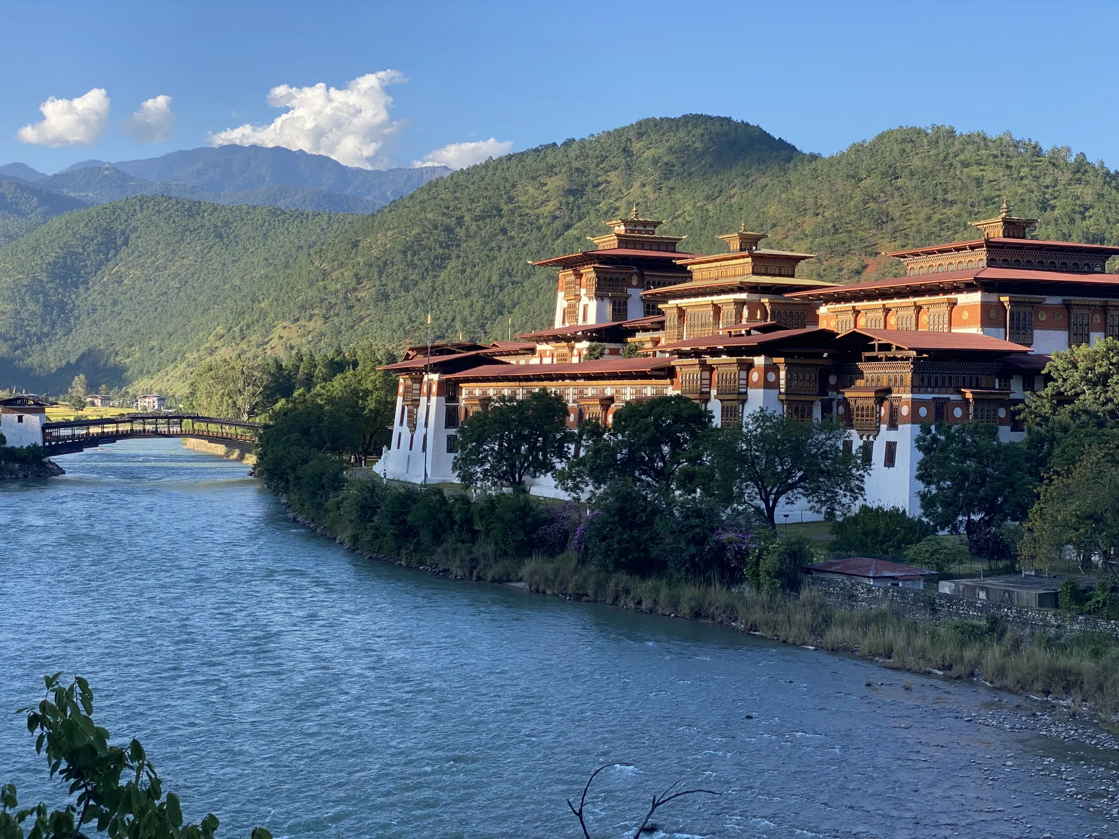 The grand Punakha Dzong fortress situated along a blue river with a wooden cantilever bridge and lush green hills in the background.