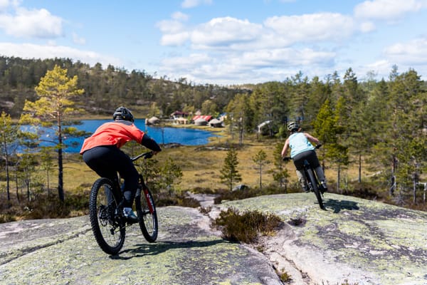 Two mountain bikers riding on a rocky trail with a lake and forest in the background.