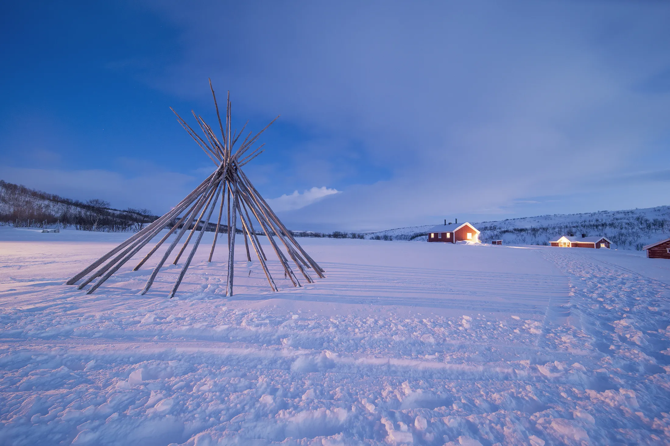 A wooden tipi frame stands in a snowy field under a blue winter sky, with illuminated houses visible in the distance.