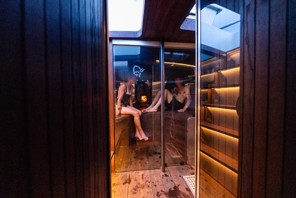 People relaxing inside a warmly lit wooden sauna seen through a glass door.