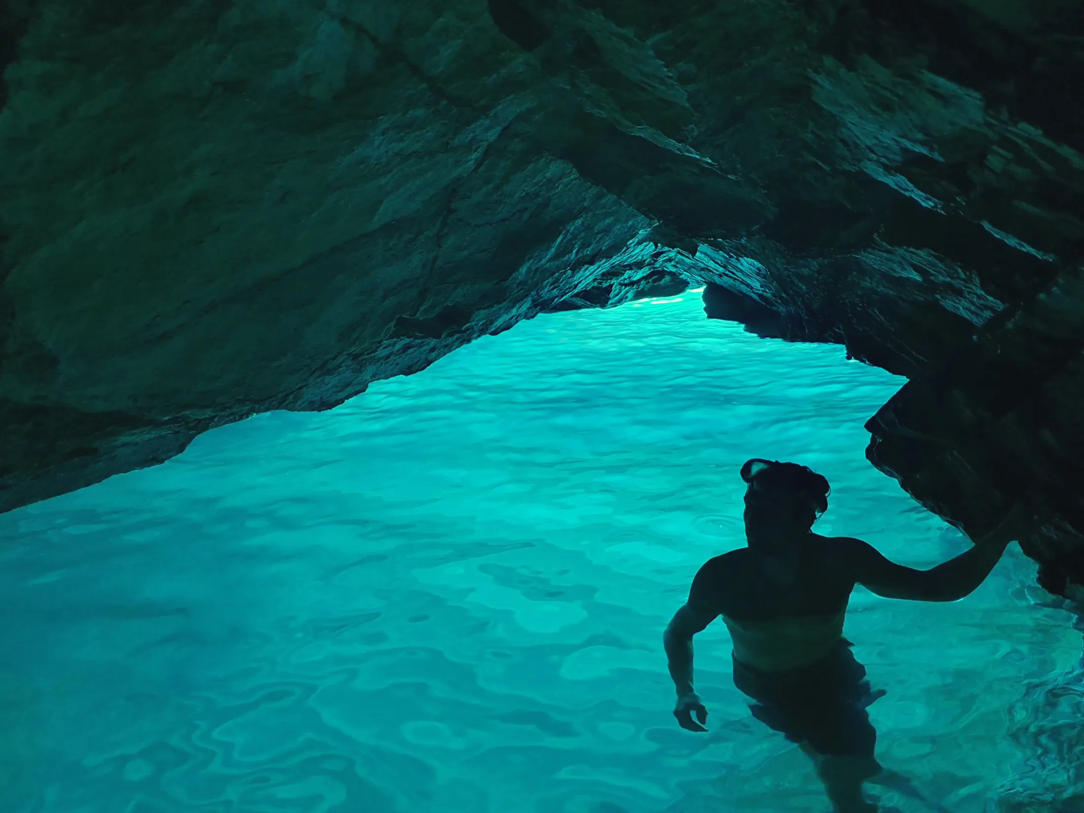 The silhouette of a snorkeler stands in glowing turquoise water inside a dark, rocky sea cave.