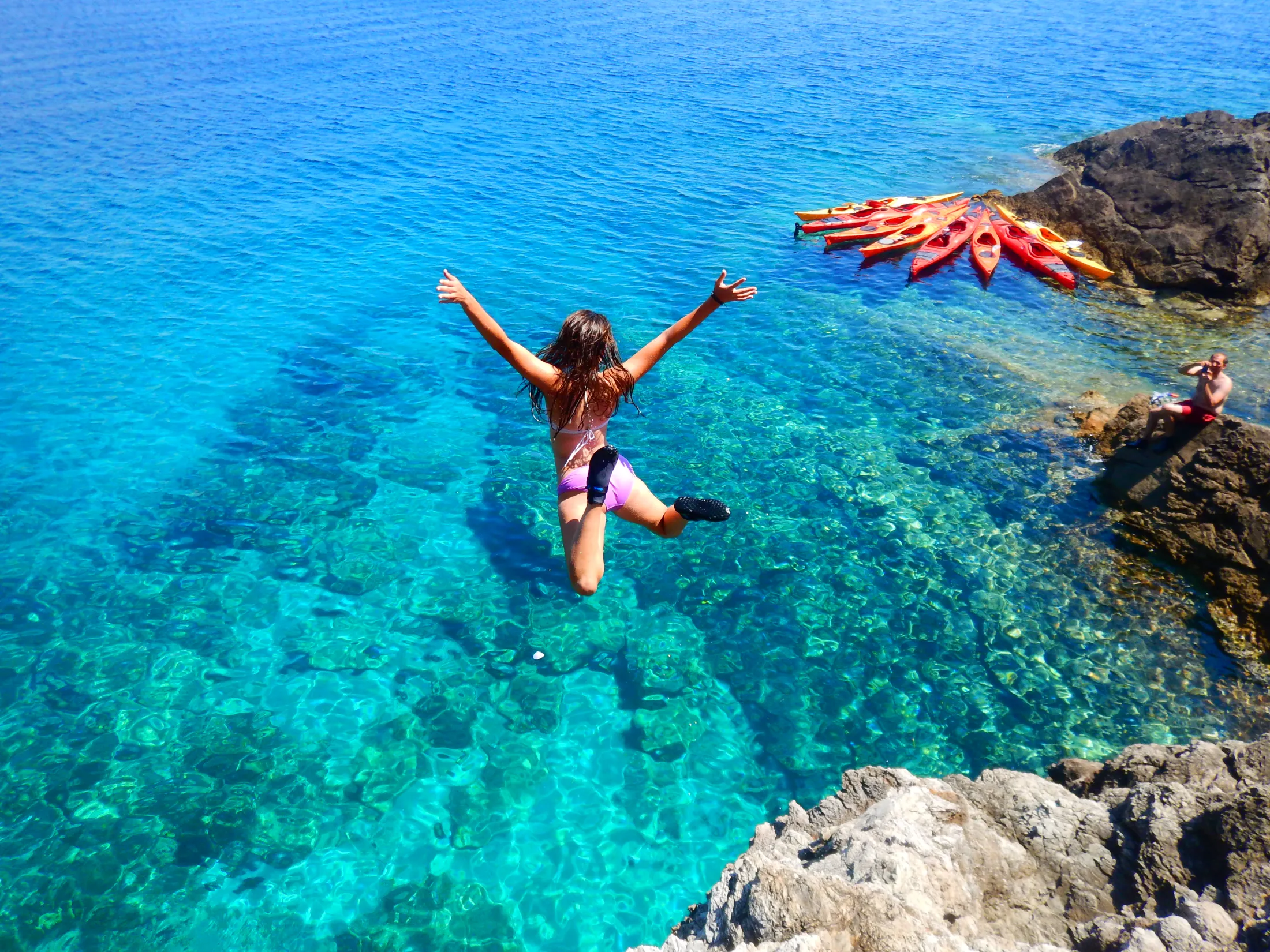A person in a bikini jumps from a rocky cliff into crystal-clear turquoise ocean water, with several colorful kayaks pulled ashore in the background.
