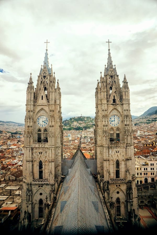 An aerial view from the Basilica del Voto Nacional in Quito, Ecuador, showing its twin neo-Gothic clock towers overlooking the city skyline under a cloudy sky.