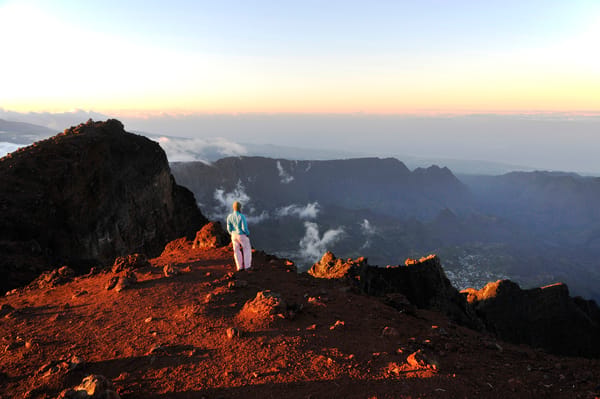 A hiker stands on a rugged, sunlit volcanic ridge looking out over a massive mountain caldera and distant valleys under a soft sunrise sky.
